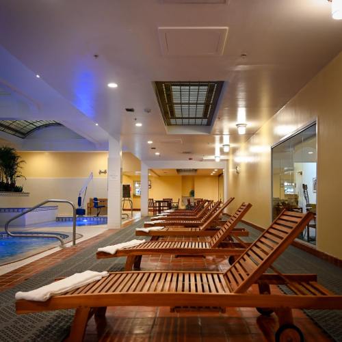 A serene indoor pool area with wooden lounge chairs, bright lighting, and plants, creating a relaxing atmosphere.