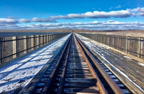 A view of railway tracks stretching into the distance, flanked by a wooden walkway and a clear blue sky with clouds.
