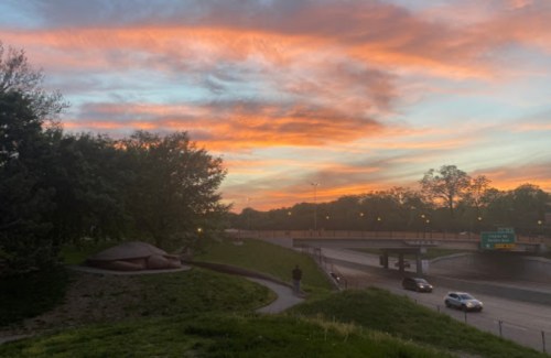 A vibrant sunset with orange and pink hues over a highway, surrounded by trees and grassy areas.