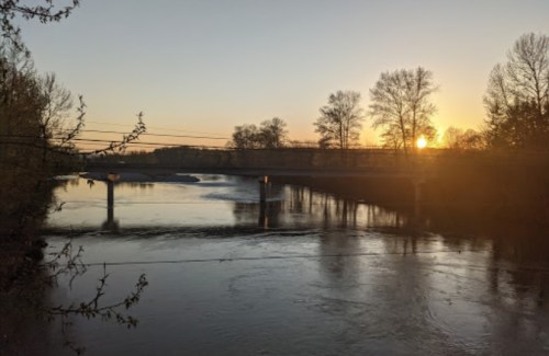 A serene river at sunset, with trees lining the banks and a bridge in the distance. The sky glows with warm colors.