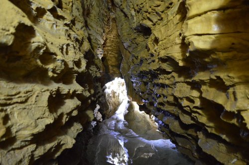 A narrow cave passage with textured yellow rock walls and light illuminating the end of the tunnel.