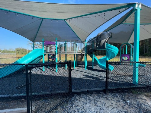 A colorful playground with slides and climbing structures, shaded by large canopies, surrounded by a fence.