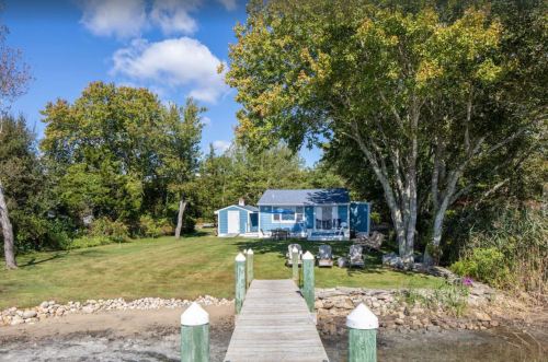 A blue cottage by the water, surrounded by trees and a grassy area, with a wooden dock leading to it.