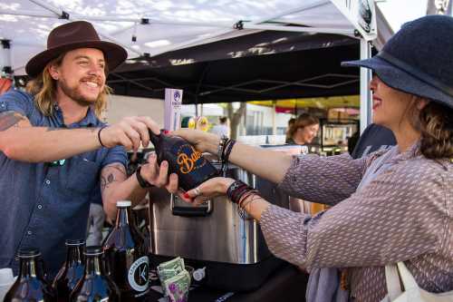 A vendor hands a woman a bottle at a market, with a cash payment visible on the table. Both are smiling.