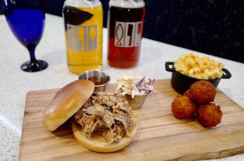 A pulled pork sandwich on a wooden board, with coleslaw, mac and cheese, and three fried balls, alongside two drinks.