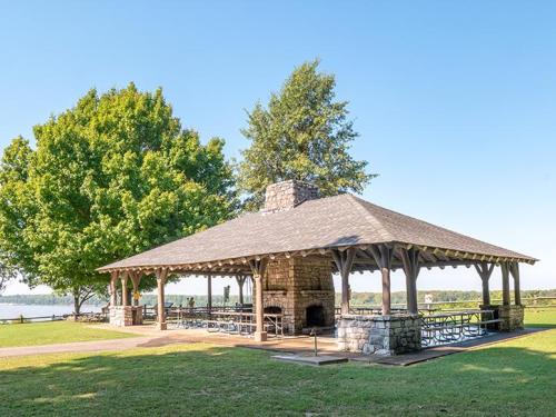 A stone pavilion with a wooden roof, surrounded by green trees and a lake in the background on a clear day.