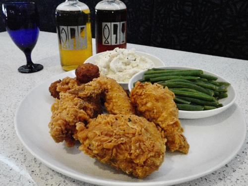 A plate of fried chicken, mashed potatoes, green beans, and a side of cornbread, with drinks in the background.