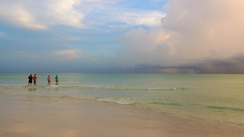A serene beach scene with four people standing in shallow water, under a cloudy sky with hints of blue.