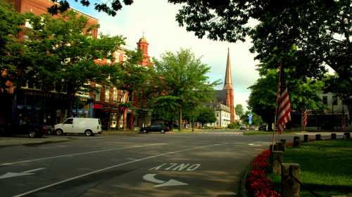 A quiet street scene with trees, buildings, and a church steeple in the background, featuring American flags and flowers.