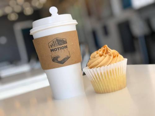 A coffee cup with a cardboard sleeve next to a frosted cupcake on a table.