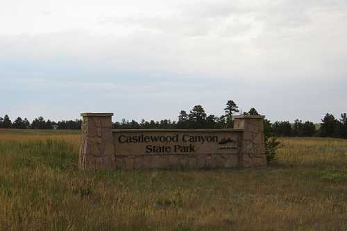 Sign for Castlewood Canyon State Park, surrounded by grassy fields and trees under a cloudy sky.