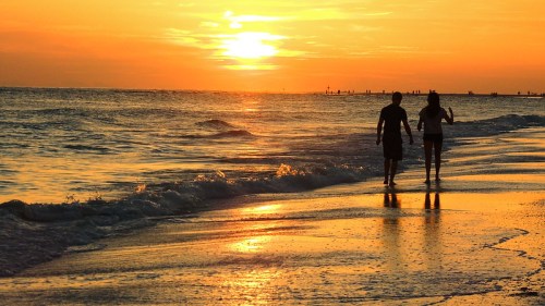 Two people walk along a beach at sunset, with golden waves reflecting the vibrant colors of the sky.