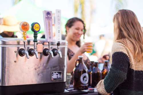 A woman at a bar taps kombucha while another woman holds a drink, with bottles and taps in the foreground.