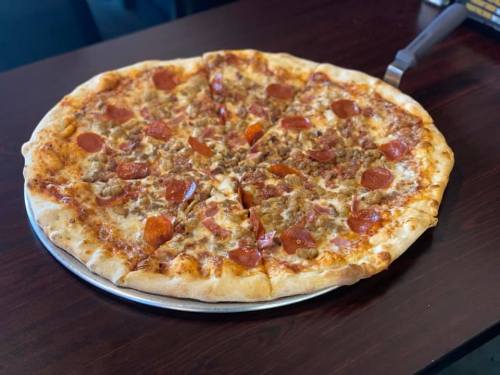 A large pizza with pepperoni and sausage, partially sliced, on a metal tray against a dark wooden table.