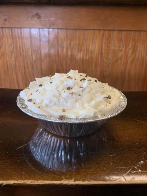 A small pie in a silver tin, topped with whipped cream and chocolate shavings, sitting on a wooden surface.