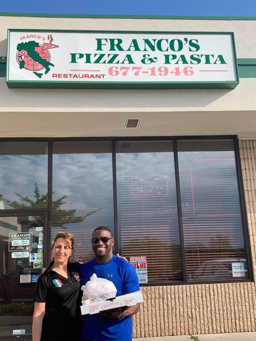 A woman and a man stand outside Franco's Pizza & Pasta, holding a pizza box, with the restaurant sign visible above.
