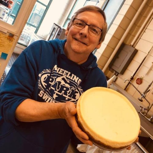 A smiling man in a blue hoodie holds a large cheesecake in a kitchen setting.