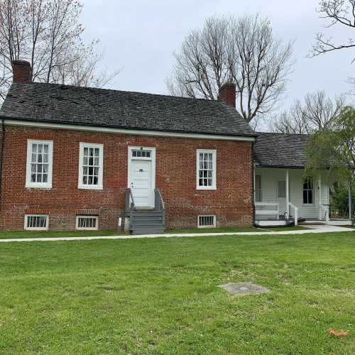 A historic brick building with a white door, surrounded by grass and trees, under a cloudy sky.