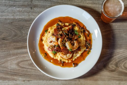A plate of creamy shrimp and grits topped with sauce and herbs, alongside a glass of beer on a wooden table.