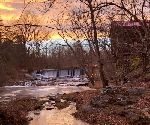 A serene landscape featuring a waterfall, a rustic building, and trees against a colorful sunset sky.