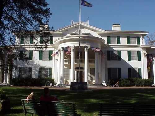 A large white mansion with green shutters, featuring a central portico and American flags, set in a grassy area.