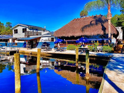 A waterfront scene featuring a thatched-roof restaurant, docked boats, and clear blue skies reflecting on the water.