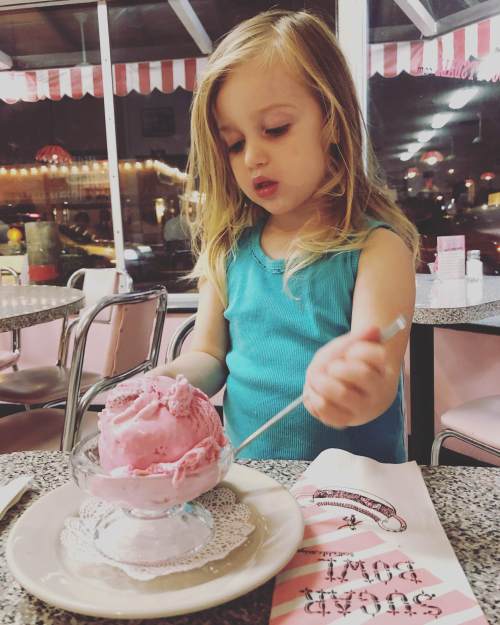 A young girl with long hair scoops pink ice cream in a retro diner, surrounded by vintage decor and soft lighting.
