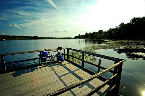 A bike with blue panniers rests on a wooden dock overlooking a calm lake surrounded by trees and a clear sky.