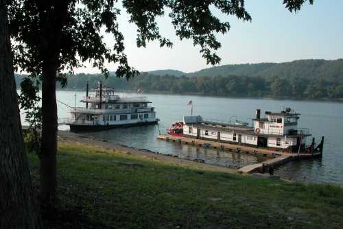 Two boats docked by a river, surrounded by trees and hills in the background under a clear sky.