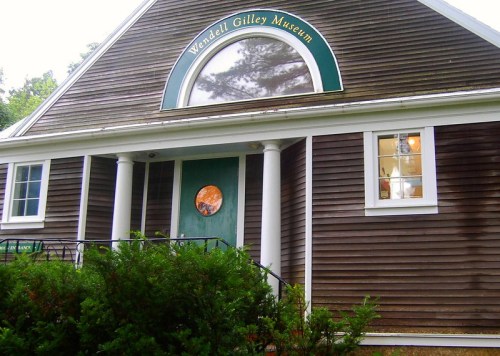 Exterior of the Wendell Gilley Museum, featuring a green door and round window, surrounded by greenery.