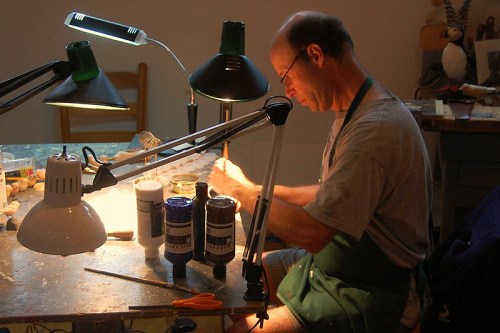 A man works at a table in a well-lit studio, surrounded by paint and tools, focused on his craft.