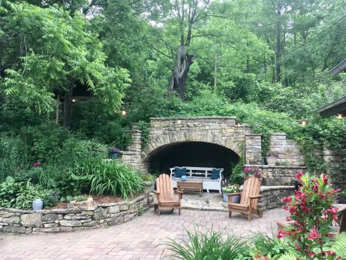 A serene garden with stone archway, lush greenery, and two wooden chairs on a brick patio.