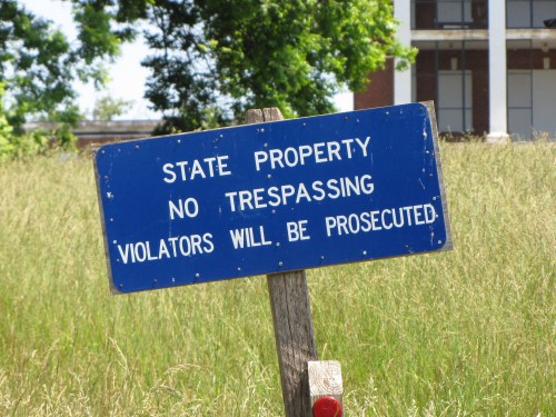 A blue sign on a wooden post reads "State Property, No Trespassing, Violators Will Be Prosecuted" in a grassy area.