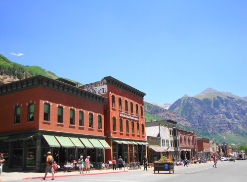 Historic buildings line a street in a mountain town, with vibrant colors and a clear blue sky in the background.