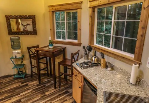 Cozy kitchen corner with a table, chairs, water cooler, and modern appliances, surrounded by large windows.