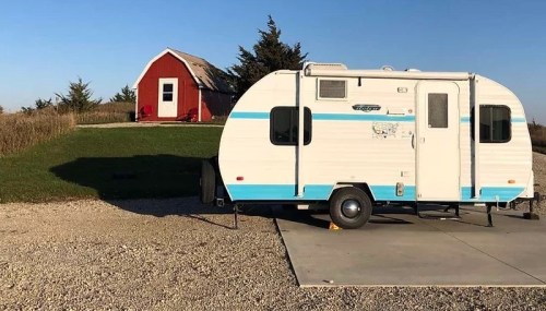 A vintage camper parked on a concrete pad next to a red barn in a rural setting with clear blue skies.