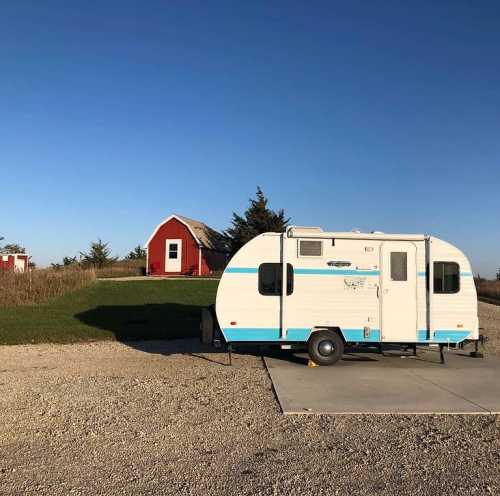 A vintage camper parked on a gravel pad, with a red cabin in the background under a clear blue sky.