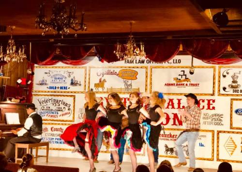 A lively stage performance with dancers in colorful costumes and a pianist, set in a vintage-themed venue.