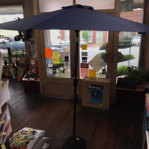 A large blue patio umbrella stands in a shop, casting shade over the wooden floor and showcasing a bright window view.