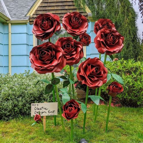 A garden featuring large, red metal roses with green leaves, alongside a sign for "Creative Design Ext."
