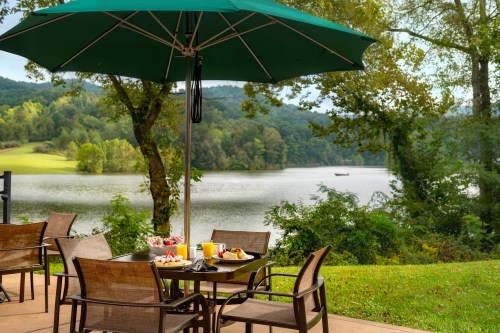 A lakeside patio with a table set for breakfast under a green umbrella, surrounded by trees and a serene view.