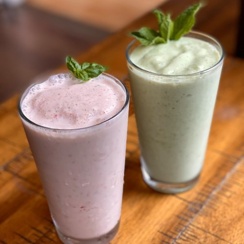 Two smoothies in glasses: one pink with a mint leaf, and one green, both on a wooden table.
