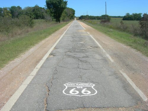 A cracked stretch of Route 66 in Oklahoma, flanked by greenery and power lines under a clear blue sky.