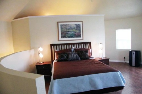 Cozy bedroom with a wooden bed, decorative pillows, lamps, and a painting on the wall, featuring warm colors and natural light.