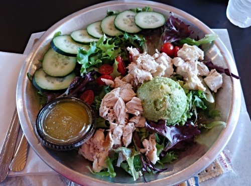 A metal plate filled with mixed greens, sliced cucumbers, cherry tomatoes, shredded chicken, and a small container of dressing.