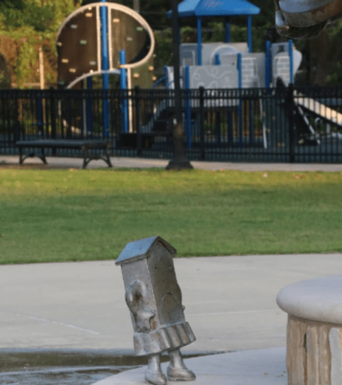 A whimsical metal sculpture of a house stands in a park, with playground equipment visible in the background.