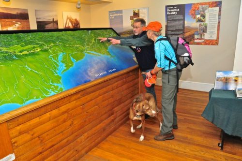 A man points at a map while a woman and a dog look on, inside a visitor center with informational displays.
