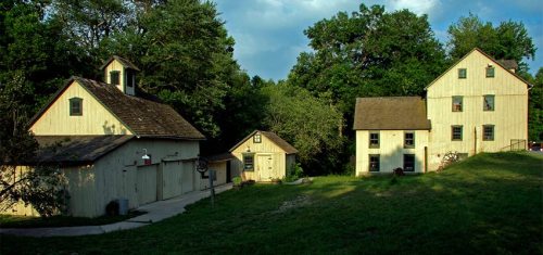 A scenic view of several rustic, light-colored buildings surrounded by lush greenery under a partly cloudy sky.