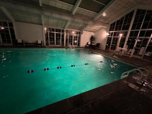 Indoor swimming pool at night, illuminated by soft lighting, with lounge chairs and large windows.
