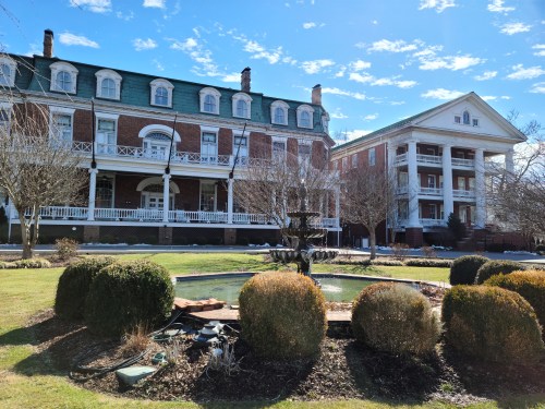 Historic buildings with porches and a fountain in a landscaped garden under a clear blue sky.